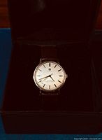 Front view of watch on black cushion in box showing silver dial, baton markers and brown leather strap