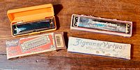 Two vintage harmonicas shown side by side with their original boxes, placed on a wooden surface. One is newer Riteho harmonica in an orange case, the other is an older Hohner harmonica labeled 'Zigeuner Virtuos' with a white box.