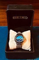 Full view of Seiko 5 wristwatch in original black Seiko box, watch displayed on white cushion pillow.