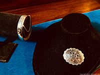 Photo showing oval shaped sterling silver brooch with floral carved decoration on black mannequin jewelry display and wide sterling silver bracelet with carved decoration and safety chain on black velvet stand.