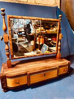 Full front view of antique dressing mirror showing wooden frame, mirror surface, three bowed front drawers, and bun feet on a blue backdrop.