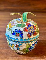 Front view of an apple-shaped trinket box showing detailed cloisonné enamel floral patterns and brass leaf and stem on top, with a wood background.