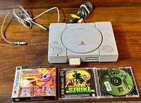 Sony PlayStation 1 console with power and AV cables on wooden table. Spyro Collector's Edition, Nuclear Strike, and Command & Conquer: Red Alert games positioned in front.