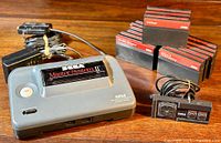 Full overview of grey Sega Master System II console, black power adapter, wired controller, and nine stacked game cartridges on wooden table.