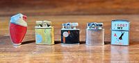 Front view of five vintage lighters of various shapes and finishes arranged side by side on wooden surface.