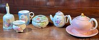 Tray lot of fine bone china including pink teapot with lid and saucer, white floral teapot, decorative creamer, sugar bowl, mug, and bell arranged on wooden surface.