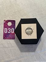 Photo showing the 1967 Canada 25 cents silver coin mounted in a white card holder and displayed on a black hexagonal base, with a purple card 030 nearby for lot identification.