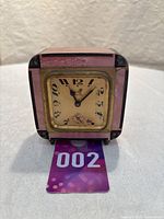 Front view of small square mantel clock with pink and black enamel frame and cream clock face showing black numbers and brass bezel.