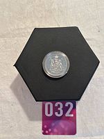 Top view of the encapsulated 1977 Canadian 50-cent Coat of Arms coin on a black hexagonal display stand with a lot card underneath.