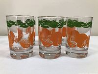 Front-facing photo showing 3 Anchor Hocking vintage orange juice glasses with orange fruit and green leaf design on clear glass.