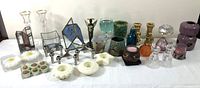 Wide view of various candle holders in glass, metal, ceramic and milk glass arranged side by side on white surface