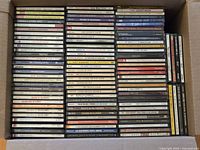 Top-down photo of a box containing multiple rows of assorted CDs, displaying title spines including classical, rock, and jazz albums.