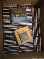 Photo showing a box of numerous CDs arranged with spines visible, featuring various artists and titles including Christmas music collections and other genres.