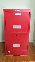 Front view of red metal filing cabinet with two drawers showing metal handles, key lock on lower drawer, and label slot on upper drawer.