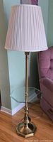Full view of the brass floor lamp with attached glass table shelf and white pleated lampshade beside a pink sofa.