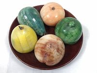 Top-down view showing six marble and onyx fruit sculptures arranged on a round wooden plate. The fruits vary in shape and color, including green, yellow, cream, and brown shades. The wooden plate is dark brown with visible wood grain.