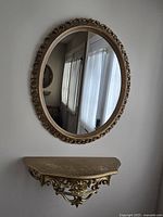 Full view of the gold oval mirror and matching ornate wall shelf mounted on a light-colored wall.