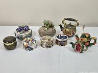 Seven trinket boxes and decorative teapots arranged on a table, showing a frog trinket box, floral boxes, and two decorative teapots.
