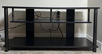 Front view of black TV stand showing three levels including a middle shelf divided into three sections, standing on tiled floor against a wall with electrical outlets and cables visible.