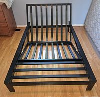 Front view of black metal twin futon frame showing slatted backrest and base, placed on wooden floor.