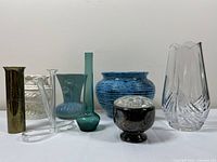 Front view of all eight assorted vases and pots arranged in a row showing varying colors and materials including glass, ceramic, crystal, and metal