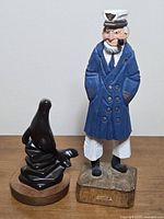 Photo showing both wooden carvings together on a plain background: the captain figurine in blue coat and the dark walnut seal sculpture on round base.