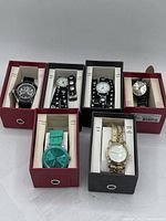 Front view of 6 ladies watches in their individual boxes showing various styles and colors including turquoise silicone, gold-tone braided strap, and black or silver-tone metal band watches
