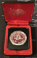 Silver dollar coin inside red and black display case with gold emblem inside lid.