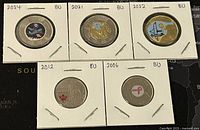 Five Canadian coins in coin holders showing colourful designs including bird, space, and awareness ribbon themes from 2004 to 2022
