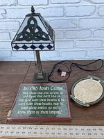 Full lot view showing the Tiffany style lamp, Irish curse plaque, and round Celtic serving tray side by side on a wooden surface with white brick background.