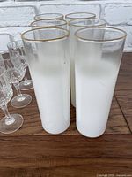 Six tall frosted white drinking glasses with gold rims arranged in two rows on wooden surface, with clear crystal-cut and brandy snifter glasses partially visible in background.