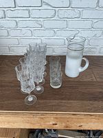 Row of 11 stemmed water glasses, 3 highball glasses and one white frosted pitcher arranged on wooden surface against white brick background