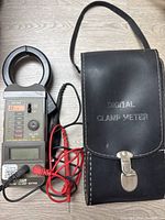 Black Extech AC/DC clamp meter with digital display, rotary dial, connected red and black test leads, and black leather carrying case.