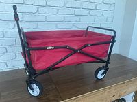Red foldable wagon with black frame and white wheels placed on wooden surface against white brick wall.
