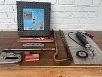 Photo showing pegboard, magnetic socket locking extension, grease gun, crowbar, hacksaw on wooden work surface against white brick wall