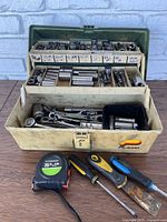 Full view of the beige Plano tackle box open showing multiple compartments with assorted sockets, wrenches, and tools inside. Three screwdrivers and a tape measure placed in front.