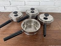 Three Echo Prestige kitchen pots displayed: two with lids, one strainer with handle; stainless steel with black handles.