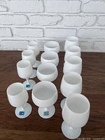 Thirteen white opaline lead crystal drinking glasses arranged on a wooden surface, varying in bowl shape and size with white stems and bases.