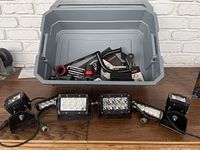 Wide photo showing six LED light housings arranged on wooden floor with gray plastic tote containing multiple installation parts behind.