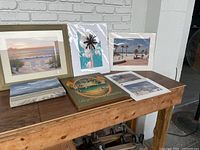 Photo of entire lot showing seven art pieces arranged on a wooden table, varied sizes and styles.