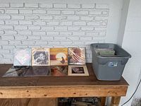 Wide shot showing a wooden table with seven vinyl albums displayed, and a gray bin containing over 75 records behind it, all sleeved.
