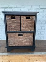 Front view of the black wooden storage side table with two small upper wicker baskets and one large lower wicker basket. The baskets are natural woven material with rectangular cutout handles.