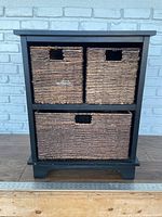 Front view of black framed storage side table with two square woven wicker baskets on the upper shelf and one larger rectangular woven wicker basket on lower shelf.