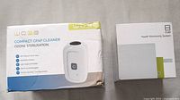 Photo of two boxed products: WOBE Compact CPAP Cleaner and Cue Health Monitoring System, both sealed in factory packaging.