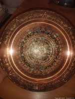 Front photo showing circular copper plate with detailed Mayan calendar motifs and colorful inlay in center and raised glyphs on rim.