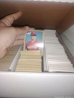 Photo showing top-down view of a cardboard box with hundreds of vintage baseball cards organized in rows, one card displayed in hand featuring a Phillies player.