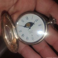 Close-up image of the watch face showing white dial with Roman numerals, moon phase display, and brand name 'Dumai Swiss Quartz'.