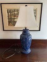 Front view of the antique blue ceramic table lamp with white lampshade placed on a wooden piece of furniture, with a framed artwork in the background.