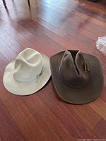 Two cowboy hats placed on wooden floor, one white with band, one brown with feather.