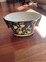 Photo of the decorative ceramic flower pot showing the orange and leaf pattern and one green handle, set on wooden floor with furniture and cushions in background.